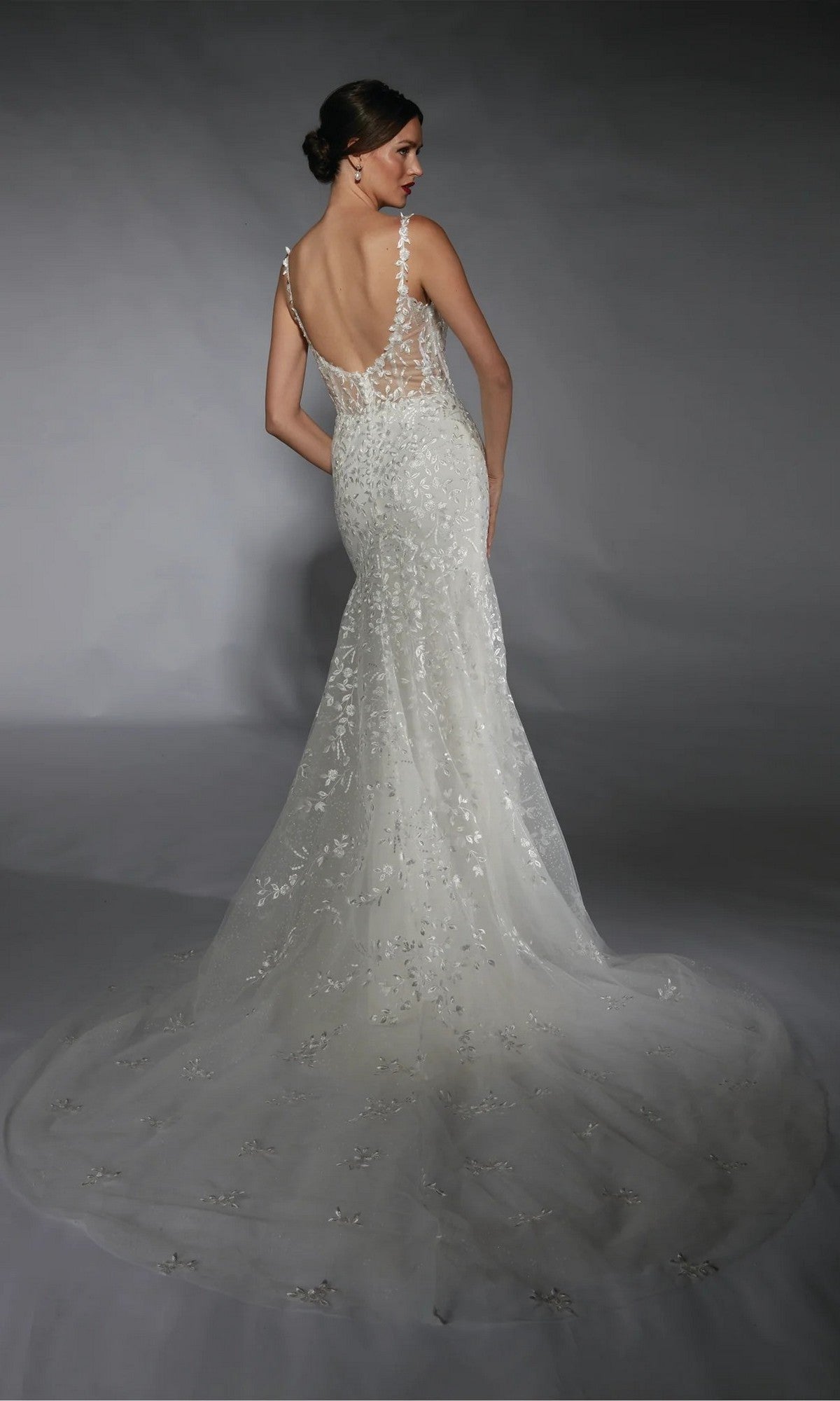 A woman in the Alyce 7120 long white dress—fitted, sleeveless, backless, with a sheer floral lace train—stands against a gray background.